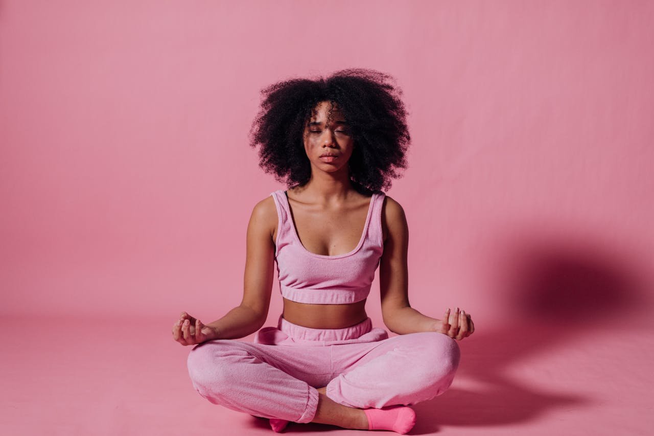 services-02 A woman with afro hair meditates in a peaceful studio with pink surroundings, embodying relaxation and zen.